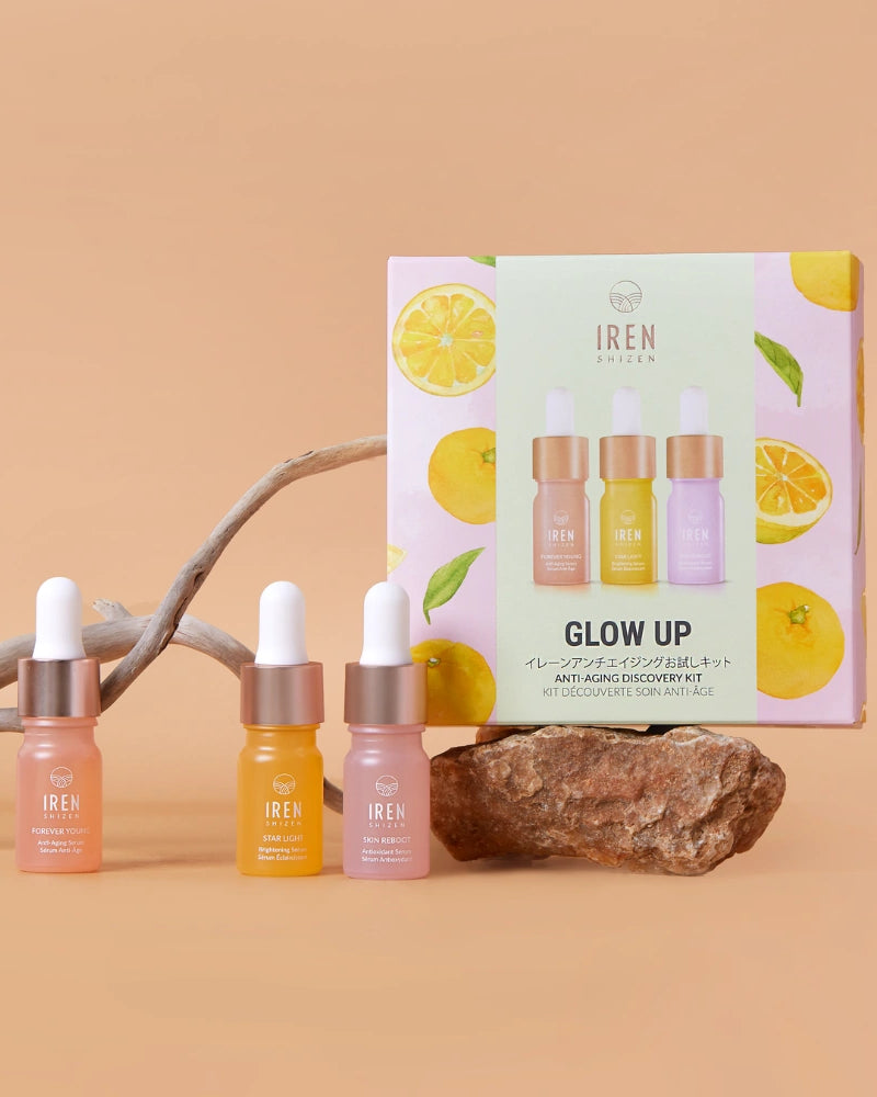 Three dropper bottles of IREN Shizen serums are shown with the GLOW UP Anti-Aging Discovery Kit box, accompanied by driftwood and citrus fruits on a beige background.