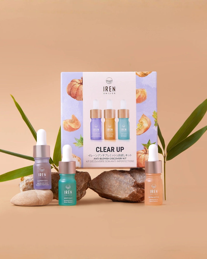 Four dropper bottles from the IREN Shizen CLEAR UP Anti-Blemish Discovery Kit, enriched with Japanese superfruits, are displayed on rocks and leaves before a kit box against a beige background.