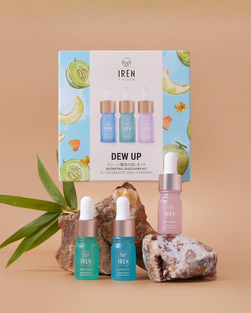 The IREN Shizen DEW UP Hydrating Discovery Kit, with three serum dropper bottles in a fruit-illustrated box, featuring antioxidant and hydrating serums for dewy skin, is displayed on rocks and a leaf against a beige background.
