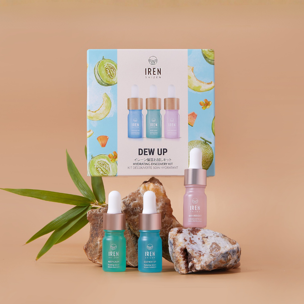 The IREN Shizen DEW UP Hydrating Discovery Kit, with three serum dropper bottles in a fruit-illustrated box, featuring antioxidant and hydrating serums for dewy skin, is displayed on rocks and a leaf against a beige background.