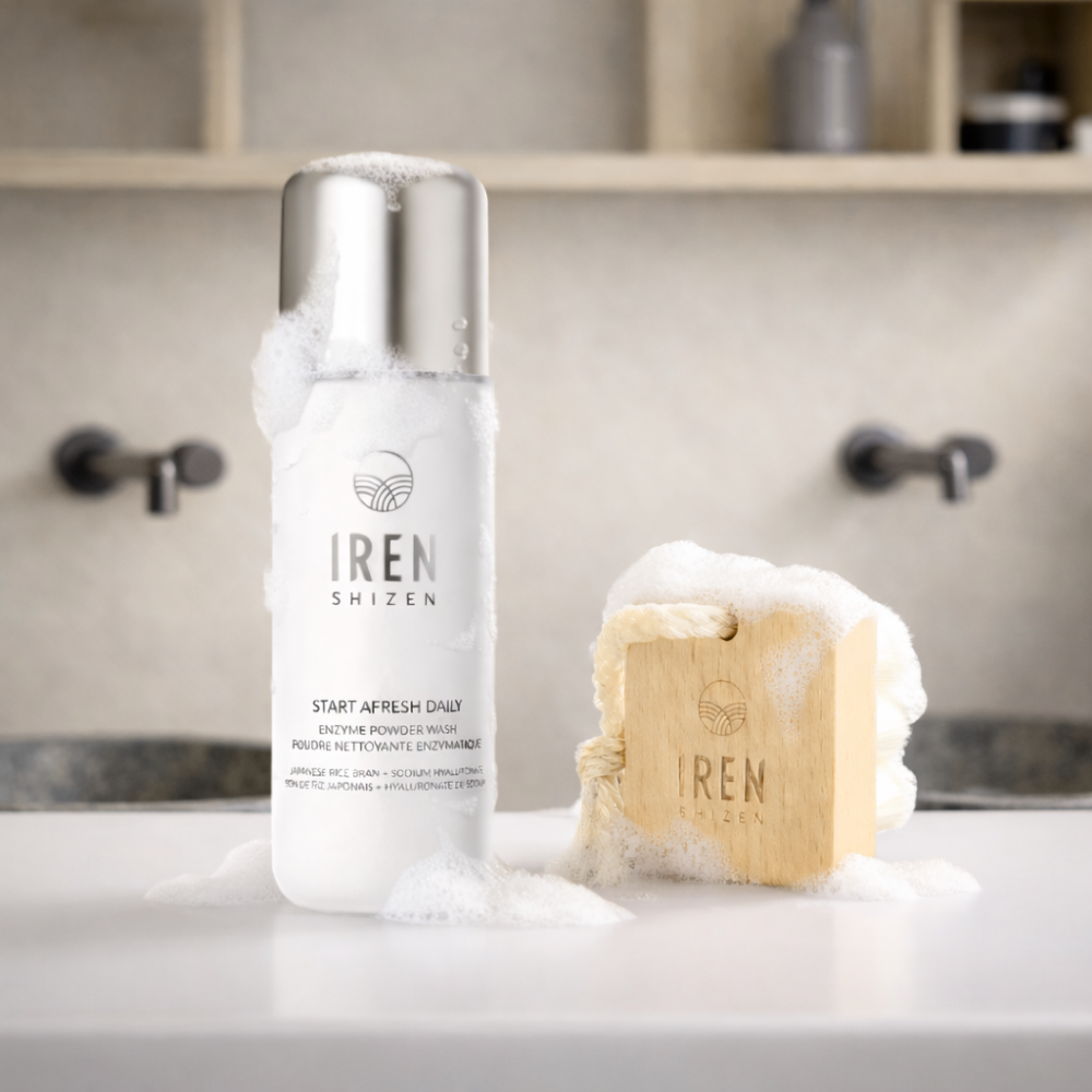 An IREN Shizen Start Afresh Daily Enzyme Powder Wash and an IREN Shizen Facial Cleansing Brush with ultra-soft bristles and foam rest on a bathroom counter, ready to offer a gentle cleanse.
