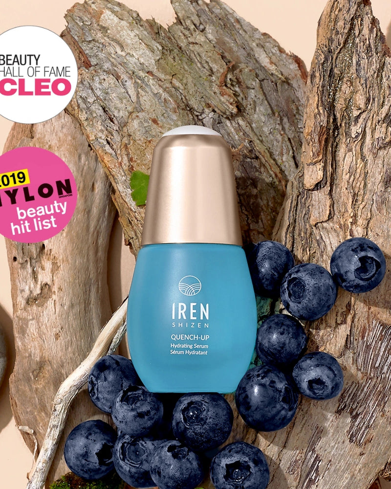 A blue bottle of IREN Shizen QUENCH-UP Hydrating Serum with hyaluronic acid is displayed on tree bark with blueberries, next to CLEO and NYLON beauty award badges.