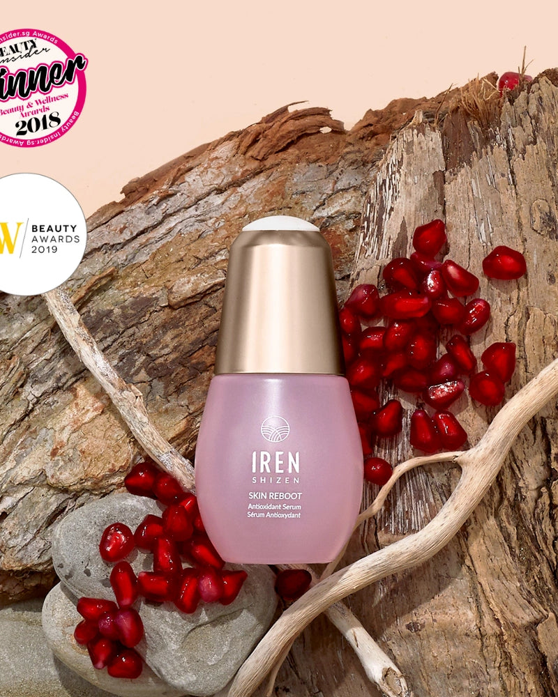 A bottle of IREN Shizen SKIN REBOOT Antioxidant Serum rests on wood with pomegranate seeds, rocks, and two beauty award badges, emphasizing its pomegranate extract for reducing dark circles. #all-groups