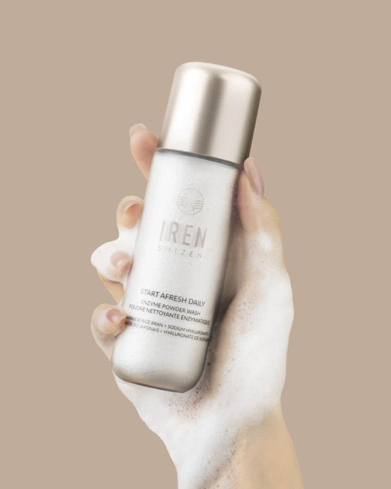 A foam-covered hand holds a silver bottle labeled "START AFRESH DAILY Enzyme Powder Wash" by IREN Shizen, a gentle rice enzyme face cleanser, against a beige background.