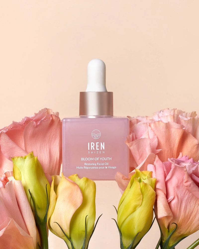 A pink dropper bottle labeled "BLOOM OF YOUTH Restoring Facial Oil" by IREN Shizen sits among pink and yellow flowers against a light beige background, highlighting its lightweight formula that supports skin barrier repair.