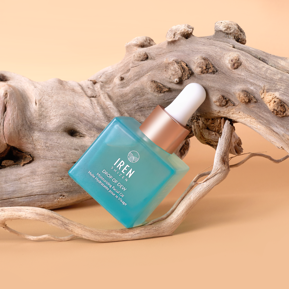 A blue glass dropper bottle labeled "IREN Shizen DROP OF DEW Moisturizing Facial Oil," a multi-tasking oil with cold-pressed oils, sits against a curved piece of driftwood on a beige background.