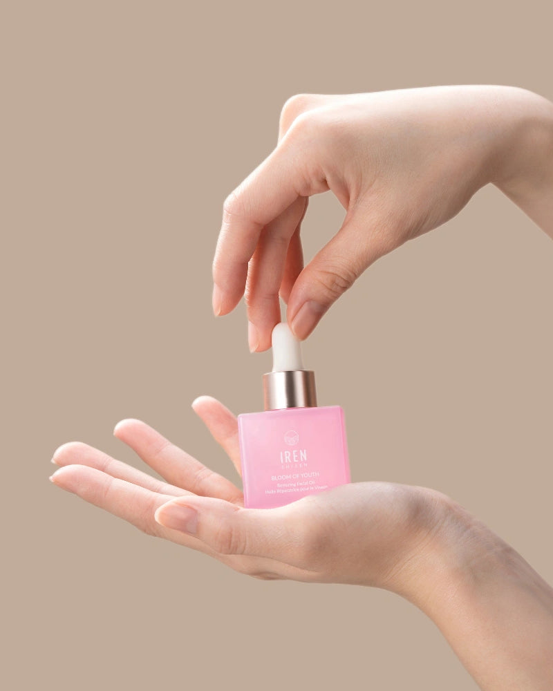 Someone holds a pink BLOOM OF YOUTH Restoring Facial Oil bottle from IREN Shizen in one hand and uses the dropper with the other against a beige background, showcasing skin barrier repair in their skincare routine.