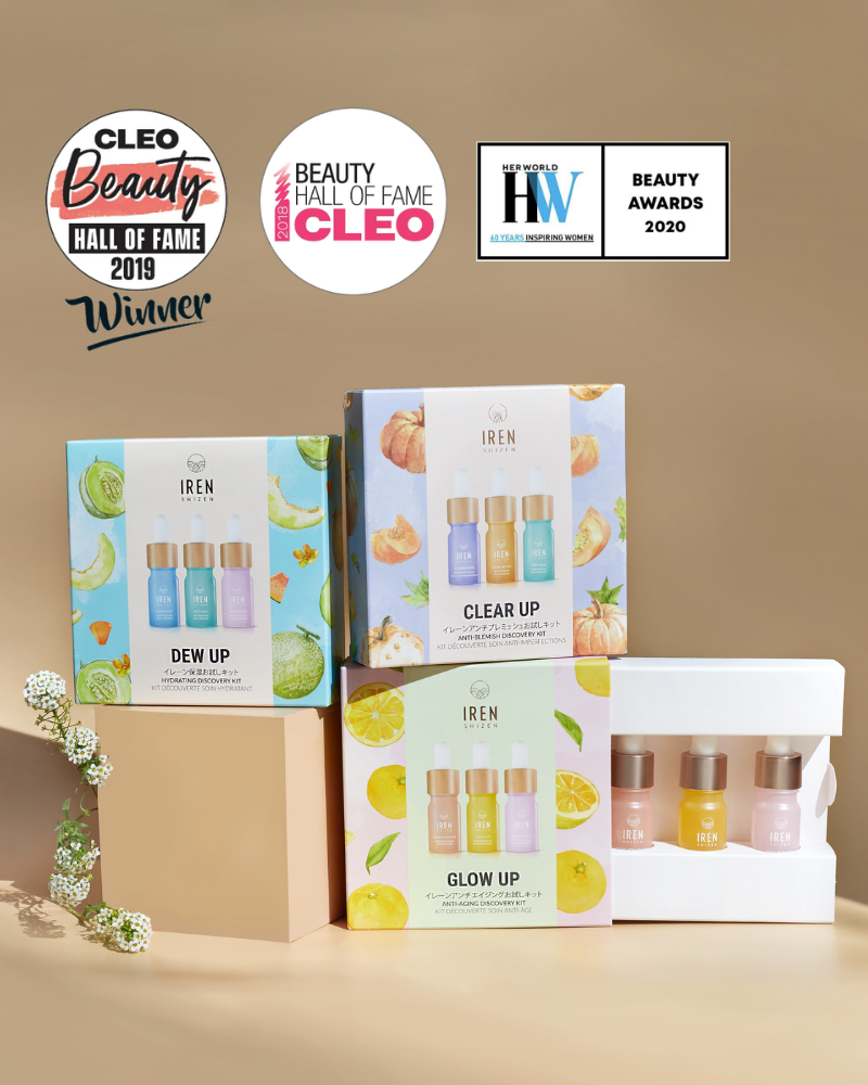Four IREN Shizen CLEAR UP Anti-Blemish Discovery Kits and three individual serum bottles are displayed on a light brown surface with shadows, featuring award badges in the background.