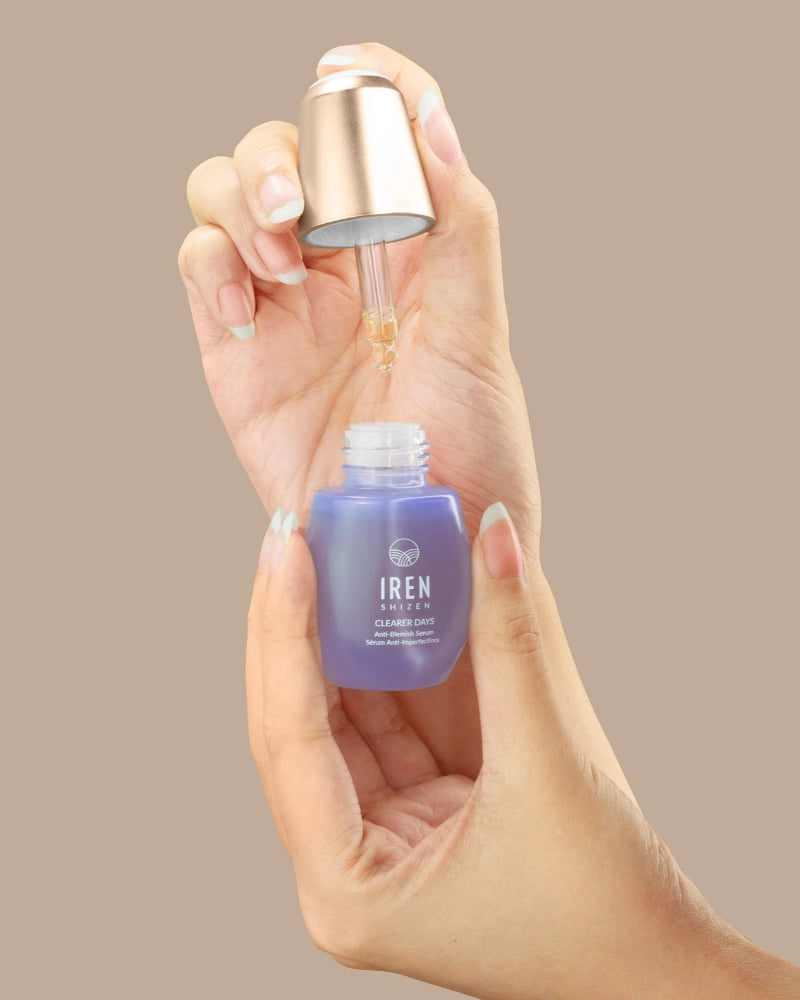 A hand holds a purple IREN Shizen CLEARER DAYS Anti-Blemish Serum bottle while another dispenses the serum with a dropper against a beige background, highlighting its acne treatment and sebum-balancing benefits. #all-groups