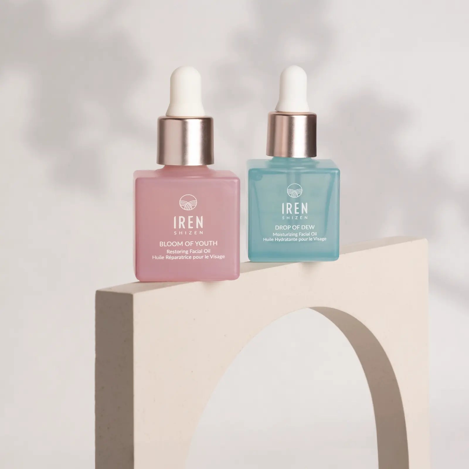 Two square IREN Shizen bottles, one pink and one blue, are displayed on a curved beige stand next to the GLOW ON THE GO Travel Light Kit, set against a neutral background.