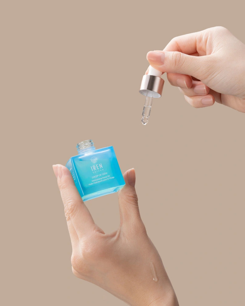 A hand holds IREN Shizen DROP OF DEW Moisturizing Facial Oil, dispensing the multi-tasking, cold-pressed oil for skin barrier repair with a dropper, set against a beige background. #all-groups