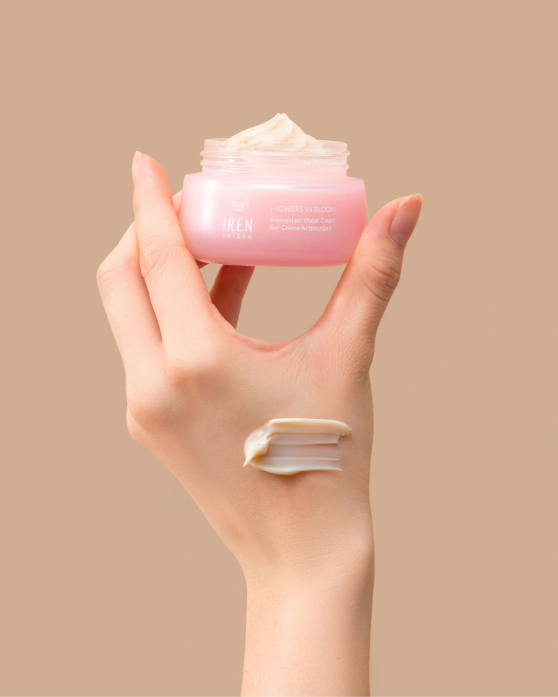 A hand holds an open jar of IREN Shizen FLOWERS IN BLOOM Antioxidant Water Cream against a beige background, with a swipe of the hydrating formula applied to the back of the hand.