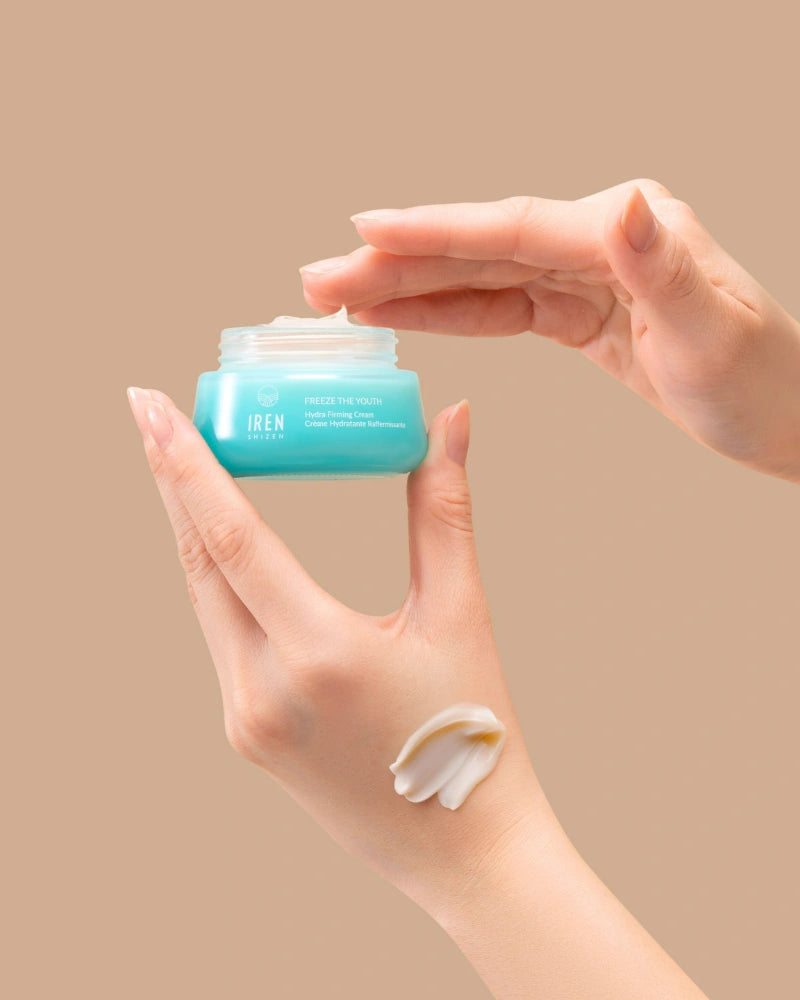 A hand holds an open jar of IREN Shizen FREEZE THE YOUTH Hydra Firming Cream while the other dips in; a smear of this hydrating, age-defying moisturizer is visible on the wrist against a beige background.