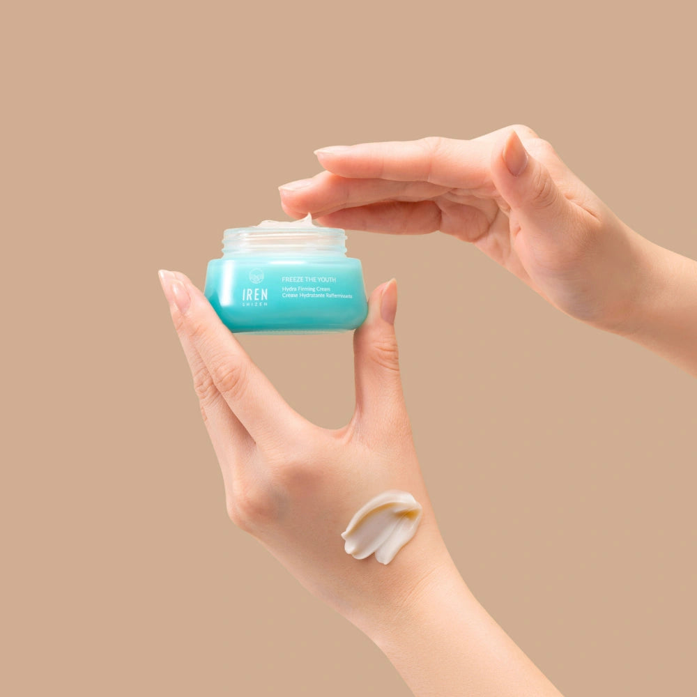 A hand holds an open jar of IREN Shizen FREEZE THE YOUTH Hydra Firming Cream while the other dips in; a smear of this hydrating, age-defying moisturizer is visible on the wrist against a beige background.
