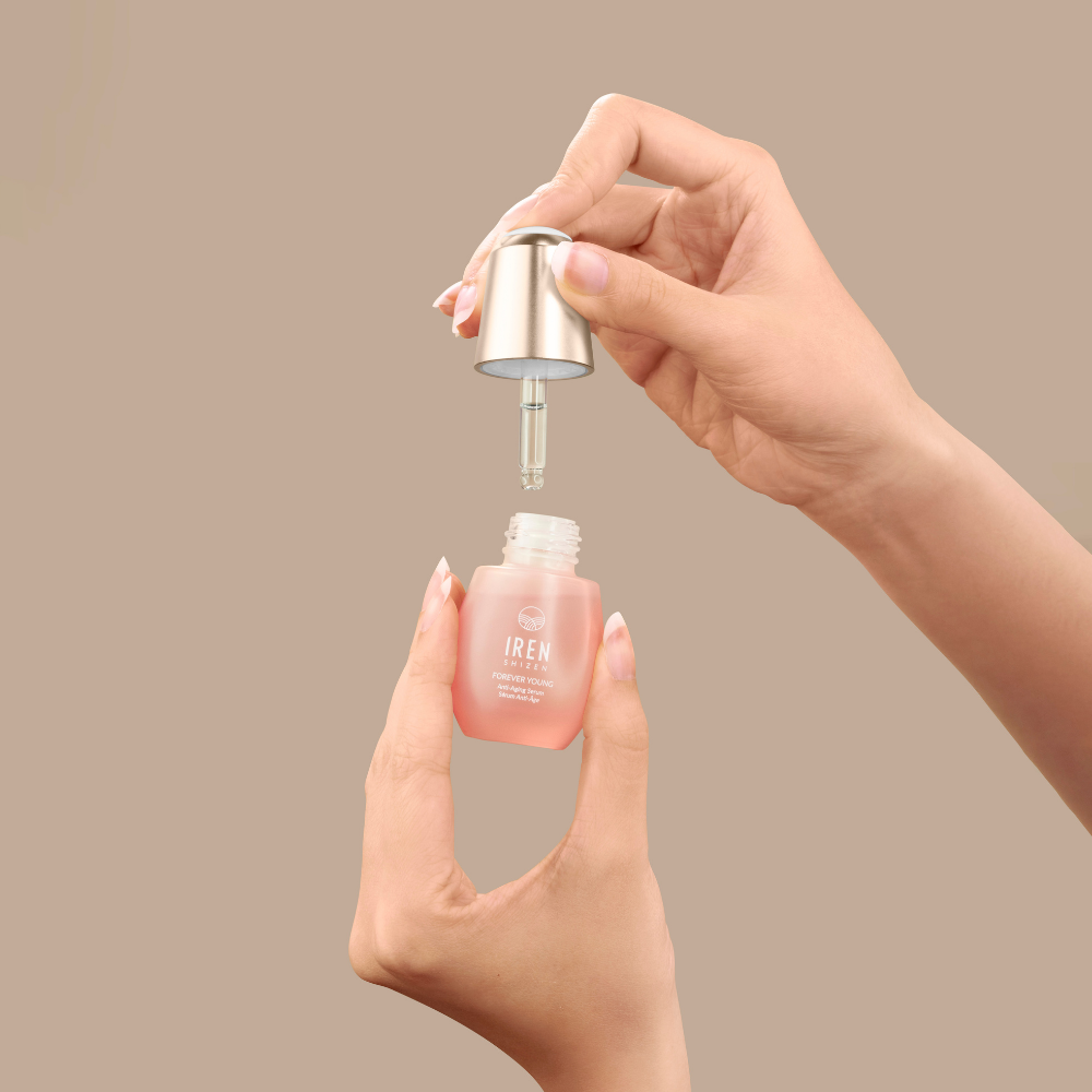 A person holds a small bottle of IREN Shizen FOREVER YOUNG Anti-Aging Serum in one hand and removes the dropper cap with the other, showcasing its anti-aging benefits against a plain beige background.