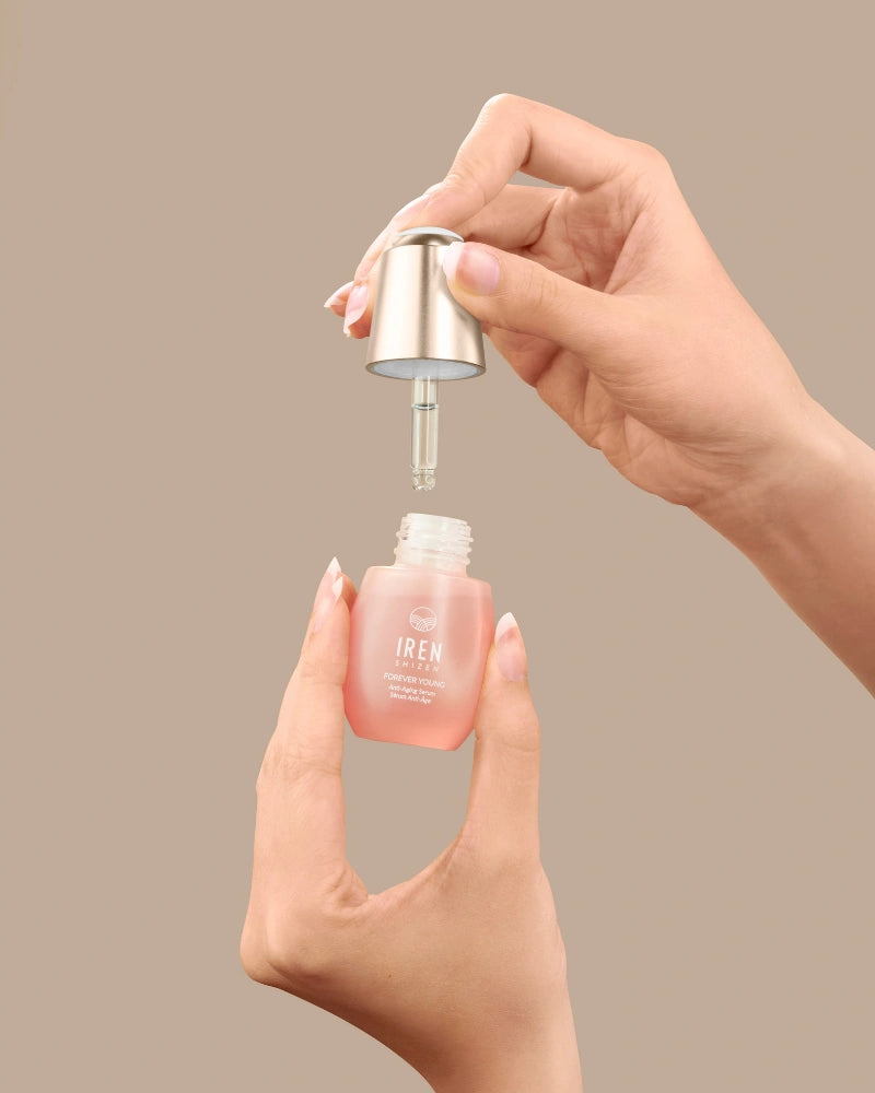 A person holds a dropper and a small glass bottle of pink FOREVER YOUNG Anti-Aging Serum by IREN Shizen, designed to support collagen production, against a plain beige background. #all-groups