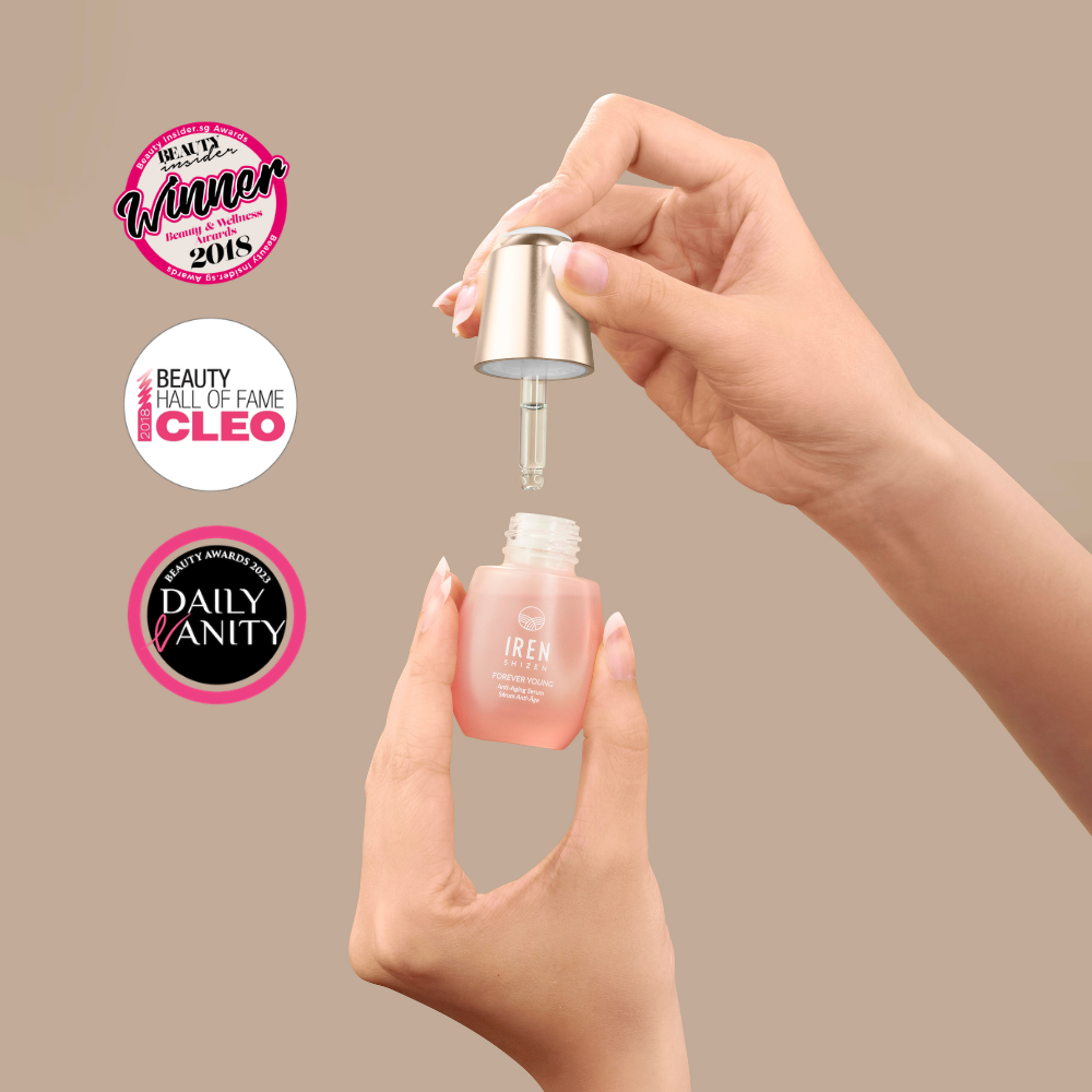 A hand holds a dropper above a small pink FOREVER YOUNG Anti-Aging Serum bottle labeled “IREN Shizen,” designed to boost collagen. Three beauty award badges appear on the left against a beige background.
