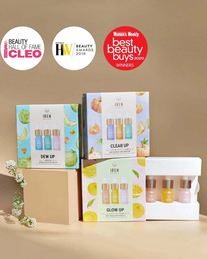 The IREN Shizen GLOW UP Anti-Aging Discovery Kit and a trio of brightening serums are displayed on a beige surface with award badges in the background.