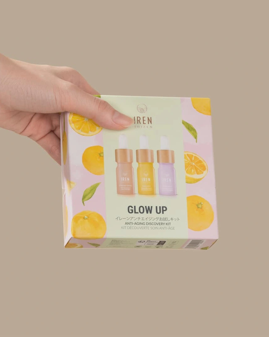 A hand holds a box of the IREN Shizen GLOW UP Anti-Aging Discovery Kit, featuring colorful citrus graphics and images of three serum bottles on the packaging.