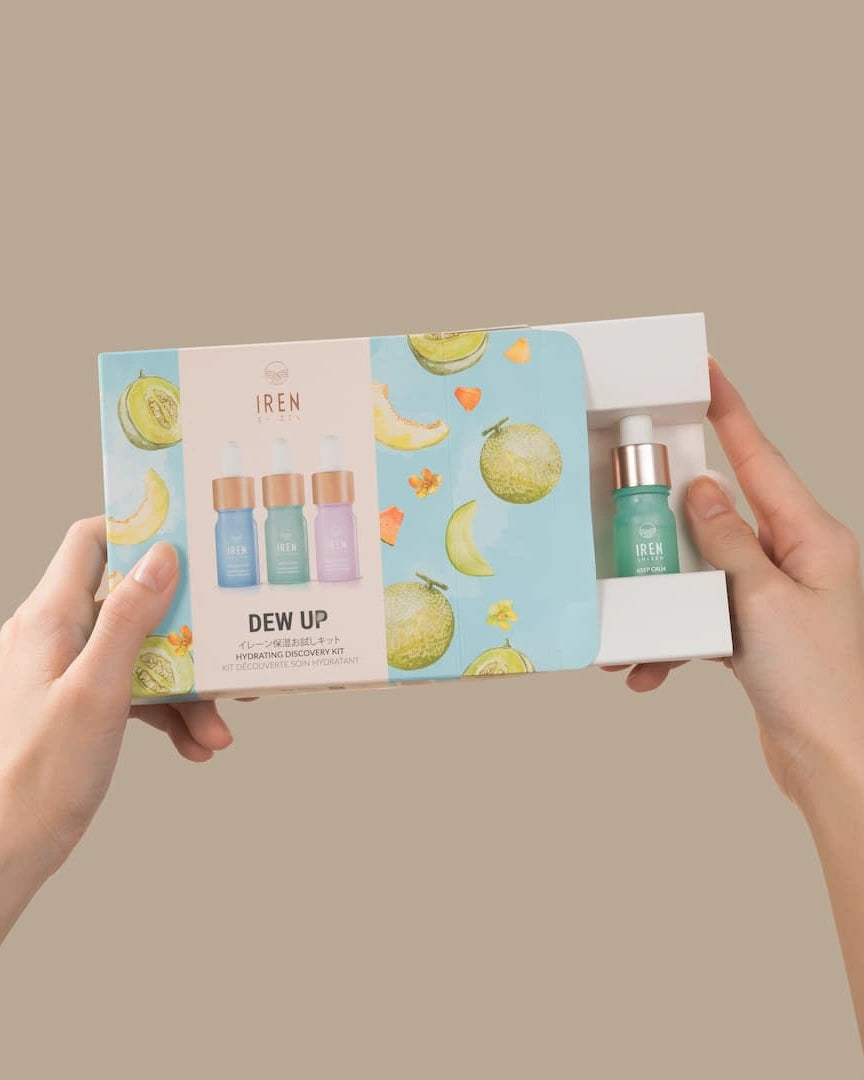 Hands hold the DEW UP Hydrating Discovery Kit by IREN Shizen, with colorful melon and flower illustrations. The partially opened box reveals a small dropper bottle of hydrating serum, perfect for achieving dewy skin.