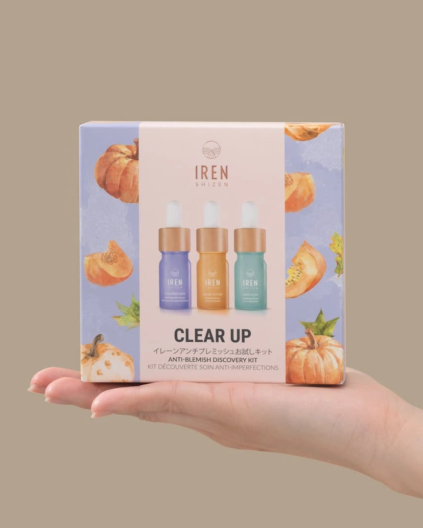 A hand holds a box of IREN Shizen CLEAR UP Anti-Blemish Discovery Kit, featuring three serums made with Japanese superfruits and decorated with pumpkins on a pastel background.