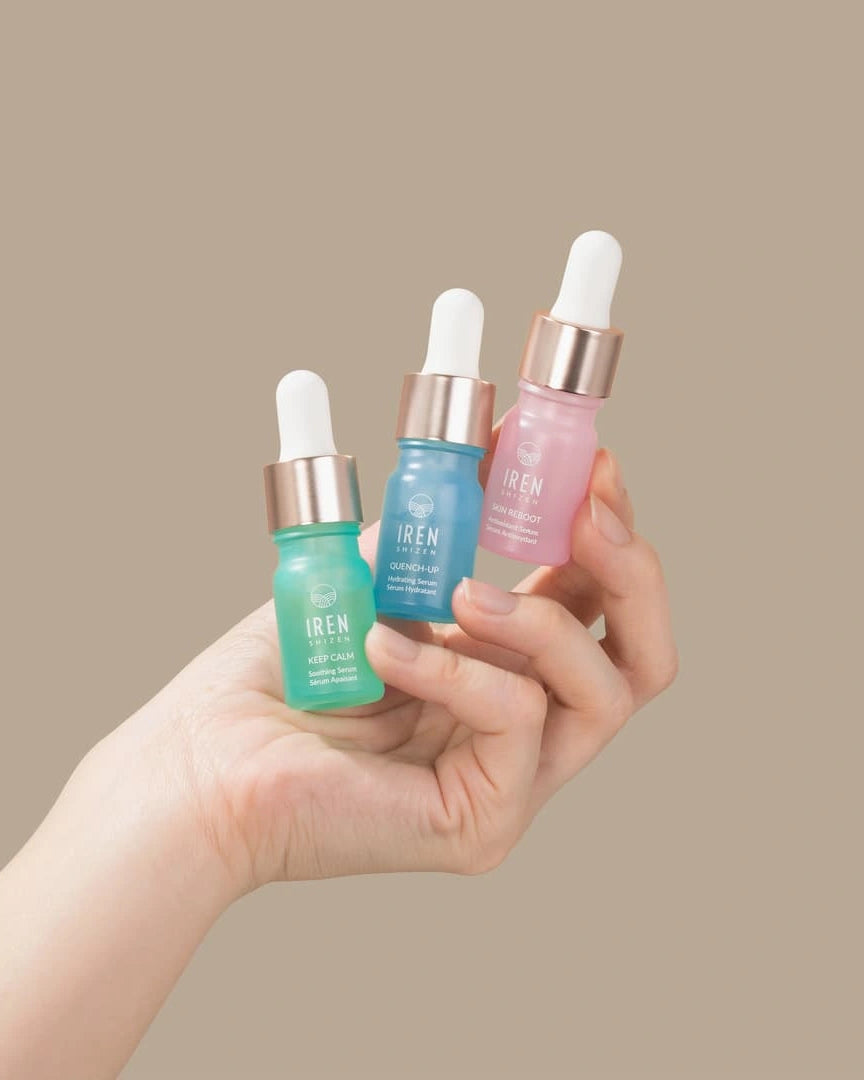 A hand holds three dropper bottles from the IREN Shizen DEW UP Hydrating Discovery Kit in green, blue, and pink, set against a plain beige background.