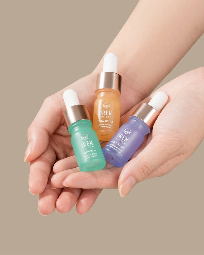 A pair of hands holds three dropper bottles from the CLEAR UP Anti-Blemish Discovery Kit by IREN Shizen, featuring serums in green, orange, and purple—each infused with Japanese superfruits—against a neutral background.