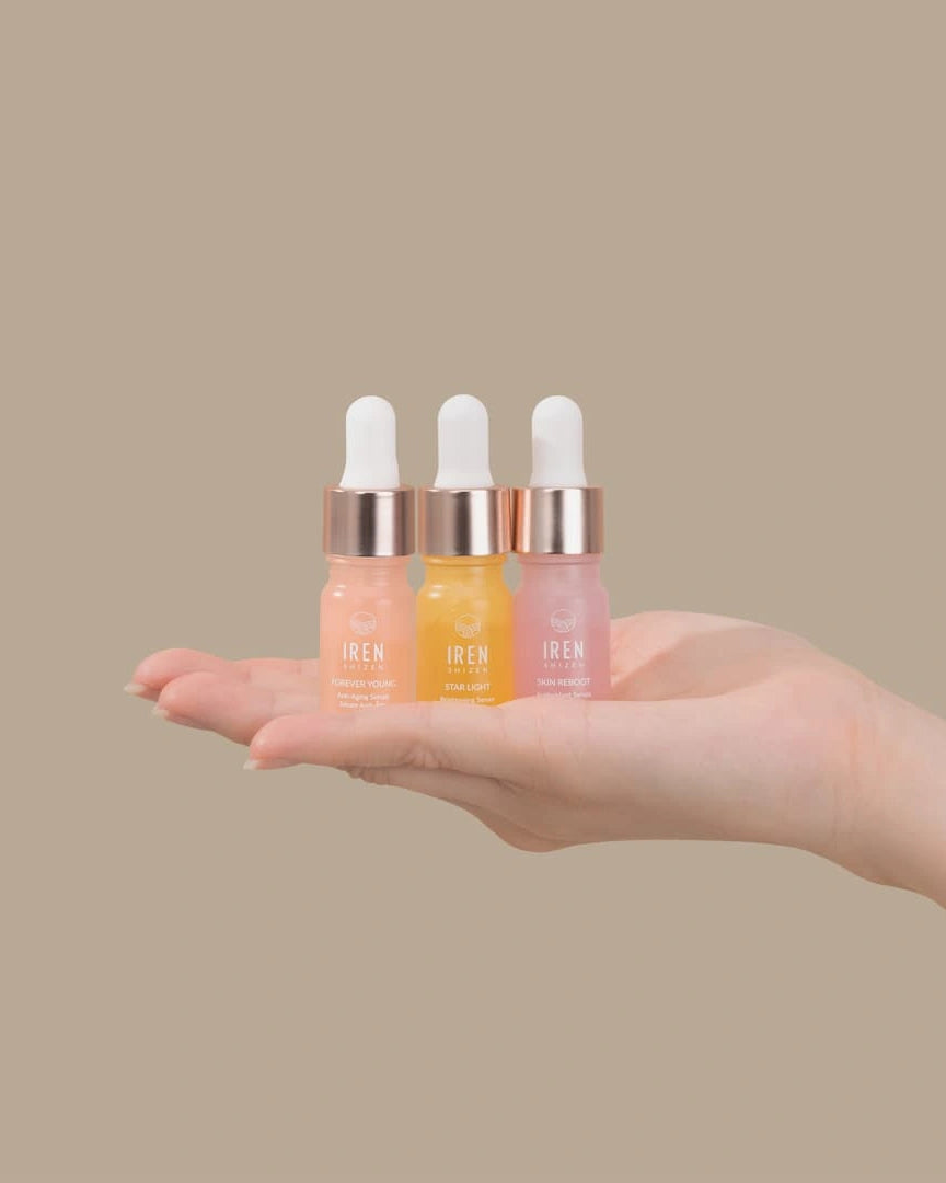 A hand holds three small dropper bottles from the IREN Shizen GLOW UP Anti-Aging Discovery Kit, featuring different colored serums against a plain beige background.