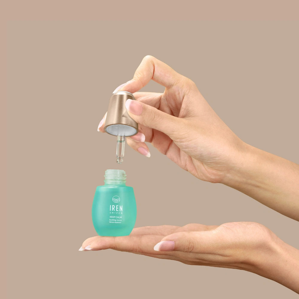 A person dispenses KEEP CALM Soothing Serum by IREN Shizen from a dropper bottle onto their palm against a plain beige background.