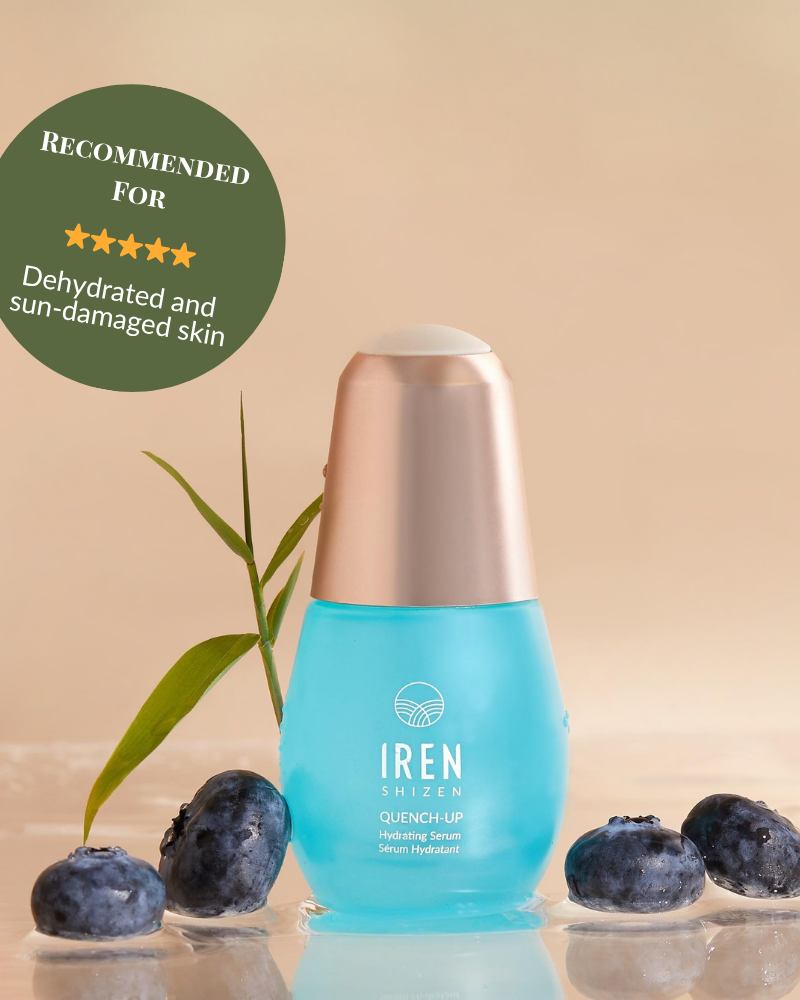 A blue bottle of IREN Shizen QUENCH-UP Hydrating Serum with hyaluronic acid sits on a wet surface among blueberries and a green leaf, labeled for dehydrated and sun-damaged skin to help achieve plump skin. #show-lang-en