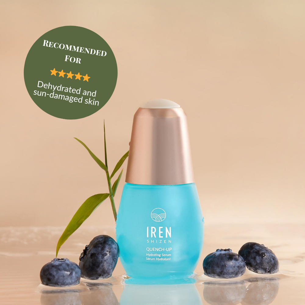 A blue bottle of IREN Shizen QUENCH-UP Hydrating Serum with hyaluronic acid sits on a wet surface among blueberries and a green leaf, labeled for dehydrated and sun-damaged skin to help achieve plump skin. #show-lang-en