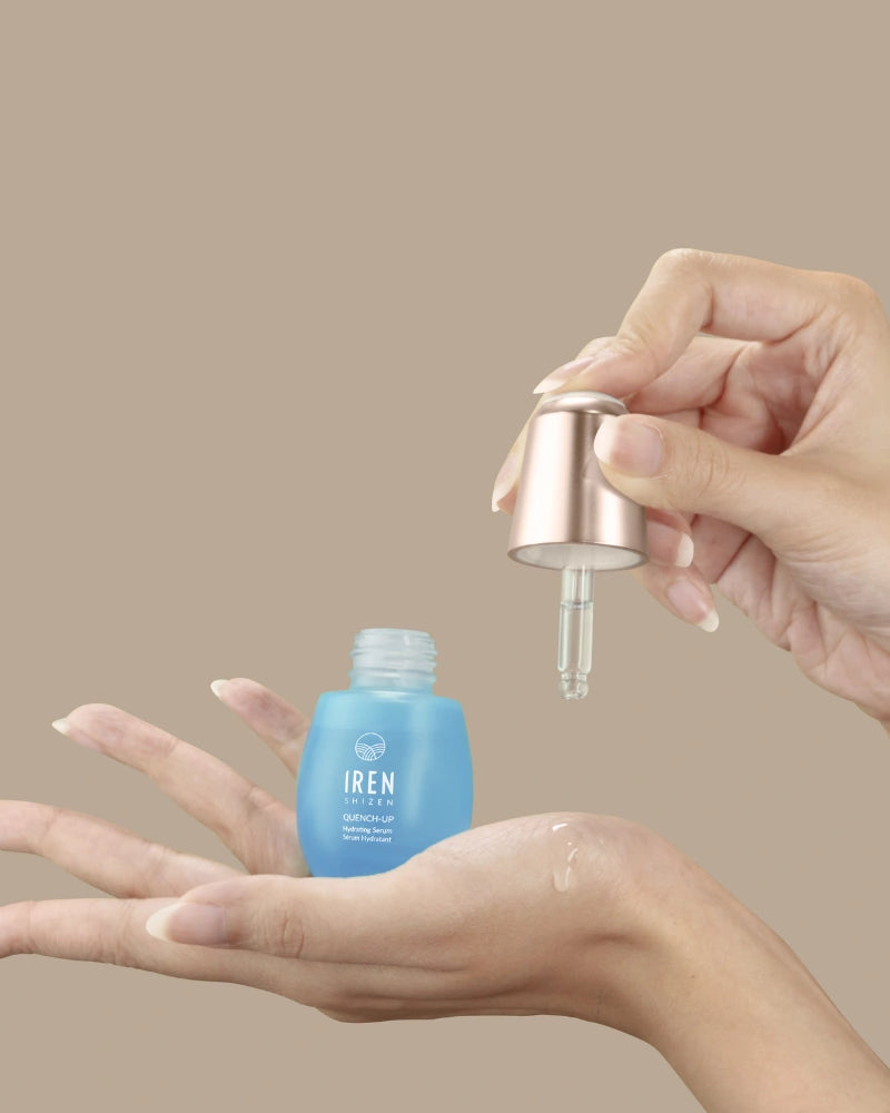A hand holds an open blue QUENCH-UP Hydrating Serum bottle by IREN Shizen while another dispenses hyaluronic acid with a dropper onto the palm, promising plump skin against a beige background.