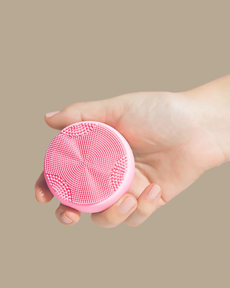 A hand holds the SKIN GENIE PRO Cleansing Brush + LED Light Therapy by IREN Shizen, featuring anti-bacterial silicone bristles, against a plain beige background.