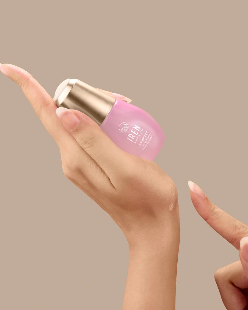 A hand holds a small pink bottle of SKIN REBOOT Antioxidant Serum by IREN Shizen with a gold cap, dispensing a drop onto the back of the hand. The serum features pomegranate extract to help reduce dark circles against a beige background. #all-groups