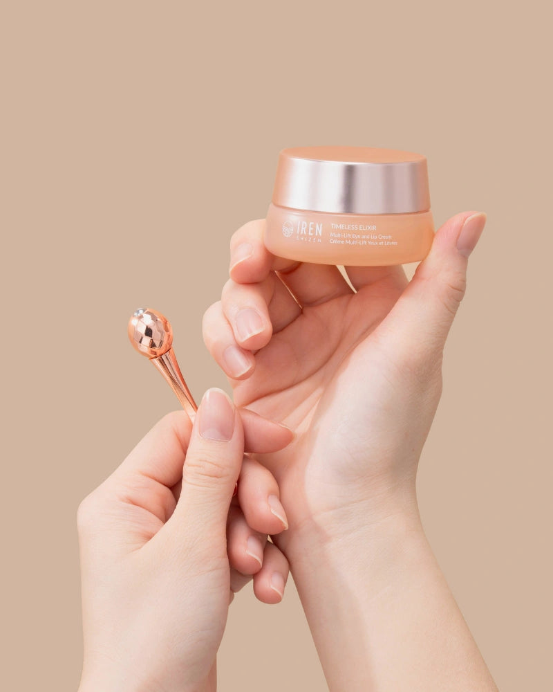 A person holds a jar of IREN Shizen TIMELESS ELIXIR Multi-Lift Eye and Lip Cream in one hand and a rose gold applicator in the other, set against a beige background—an elegant solution for treating dark circles.