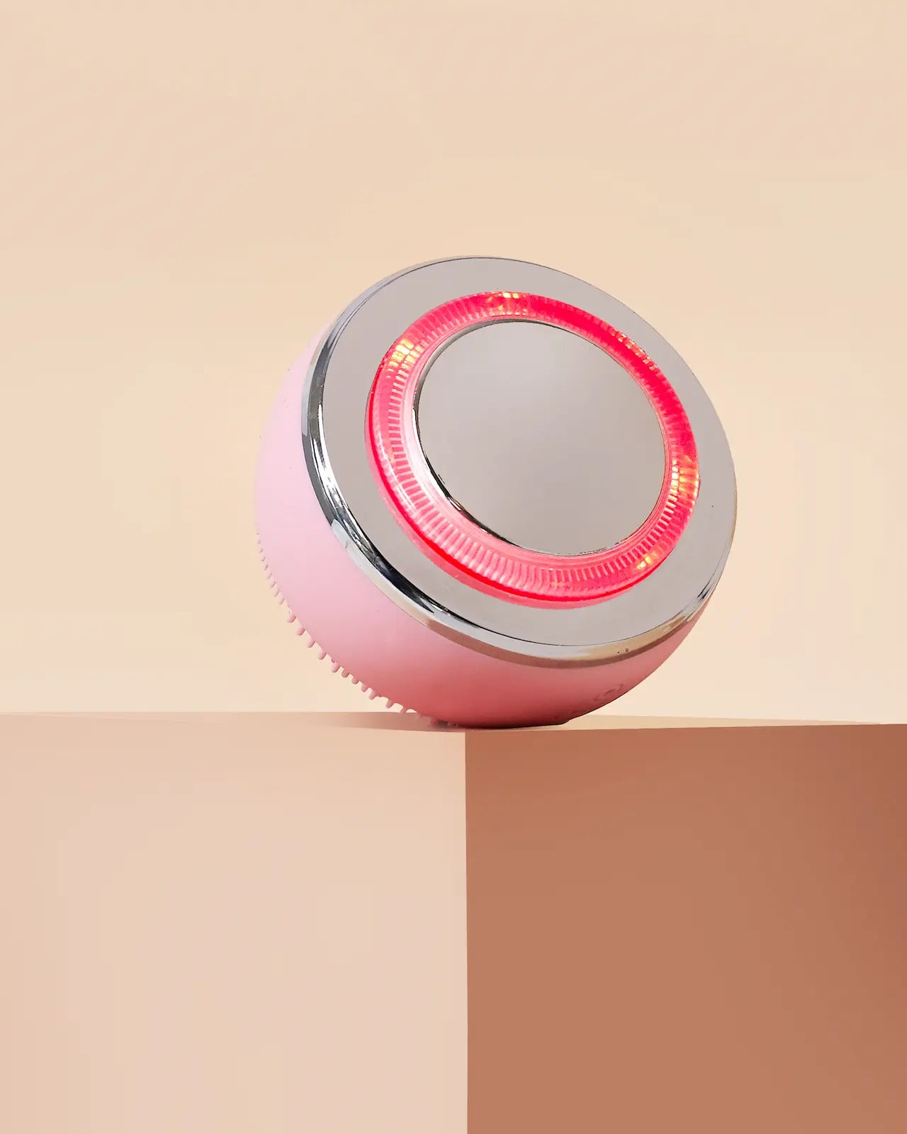 The IREN Shizen GLOW ON THE GO Travel Light Kit, a round pink and silver facial cleansing brush with red LED light therapy, rests on the edge of a beige rectangular block against a neutral background.