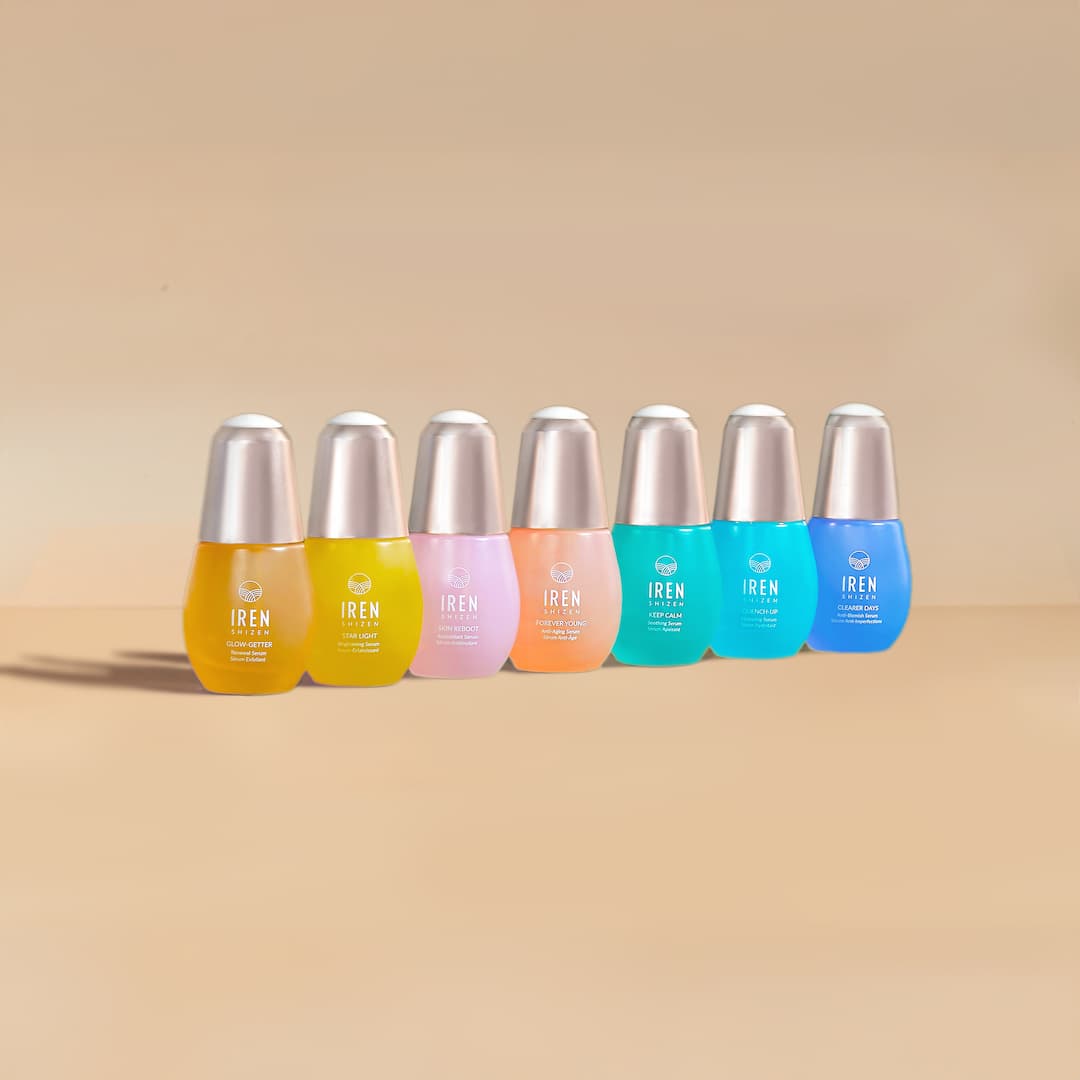 Six colorful skincare serum bottles with metallic caps arranged in a row on a beige background.
