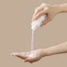 A person pours IREN Shizen’s START AFRESH DAILY Enzyme Powder Wash from a bottle onto their open palm, with a plain beige background.