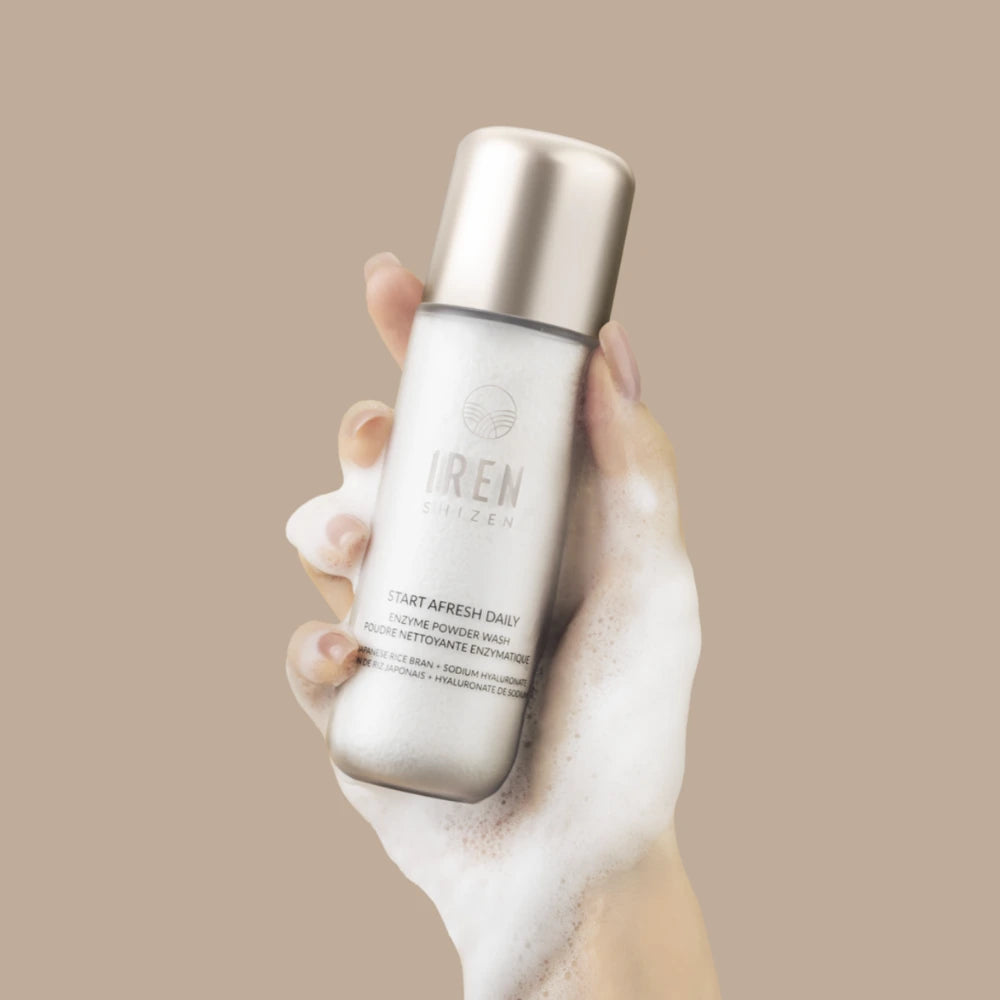 A foam-covered hand holds a silver bottle of IRÉN Shizen’s START AFRESH DAILY Enzyme Powder Wash, a gentle waterless cleansing and exfoliating powder, against a beige background.