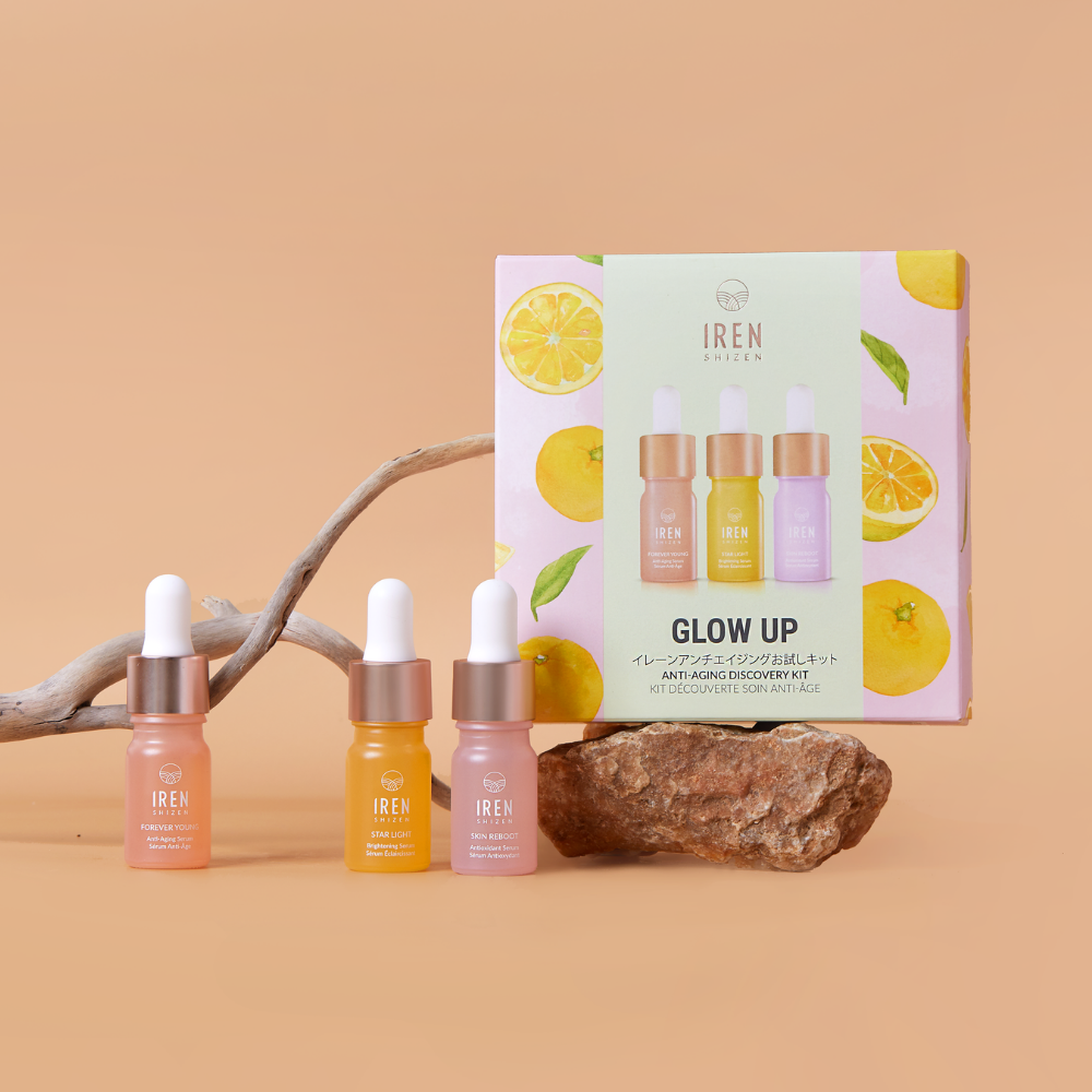 Three dropper bottles and the IREN Shizen GLOW UP Anti-Aging Discovery Kit box, featuring citrus fruit images, are displayed on a beige background with driftwood—an ideal setup to highlight your new antioxidant serum.