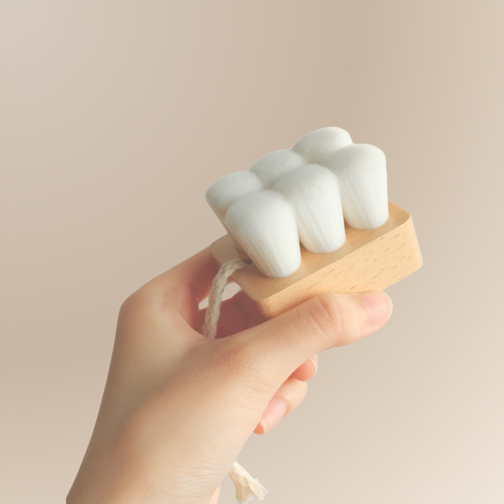 A hand holds the IREN Shizen Facial Cleansing Brush with ultra-soft, densely packed white bristles and a rope loop on the handle for gentle cleansing.