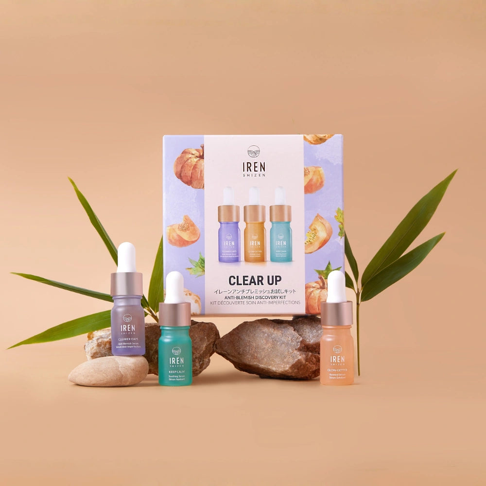 Four dropper bottles from the IREN Shizen CLEAR UP Anti-Blemish Discovery Kit, enriched with Japanese superfruits, are displayed on rocks and leaves before a kit box against a beige background.