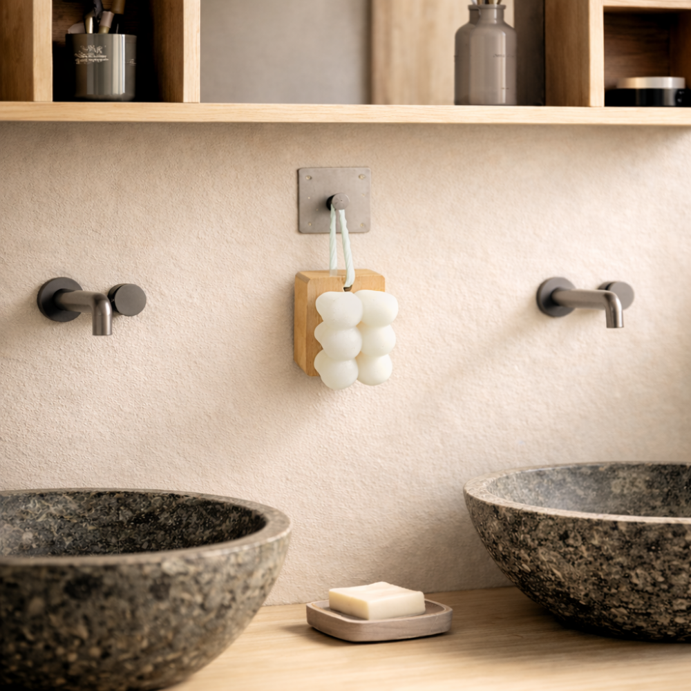 A modern bathroom with two stone sinks, wall-mounted faucets, a counter bar of soap, and the IREN Shizen Facial Cleansing Brush with ultra-soft bristles hanging on the wall between the sinks.