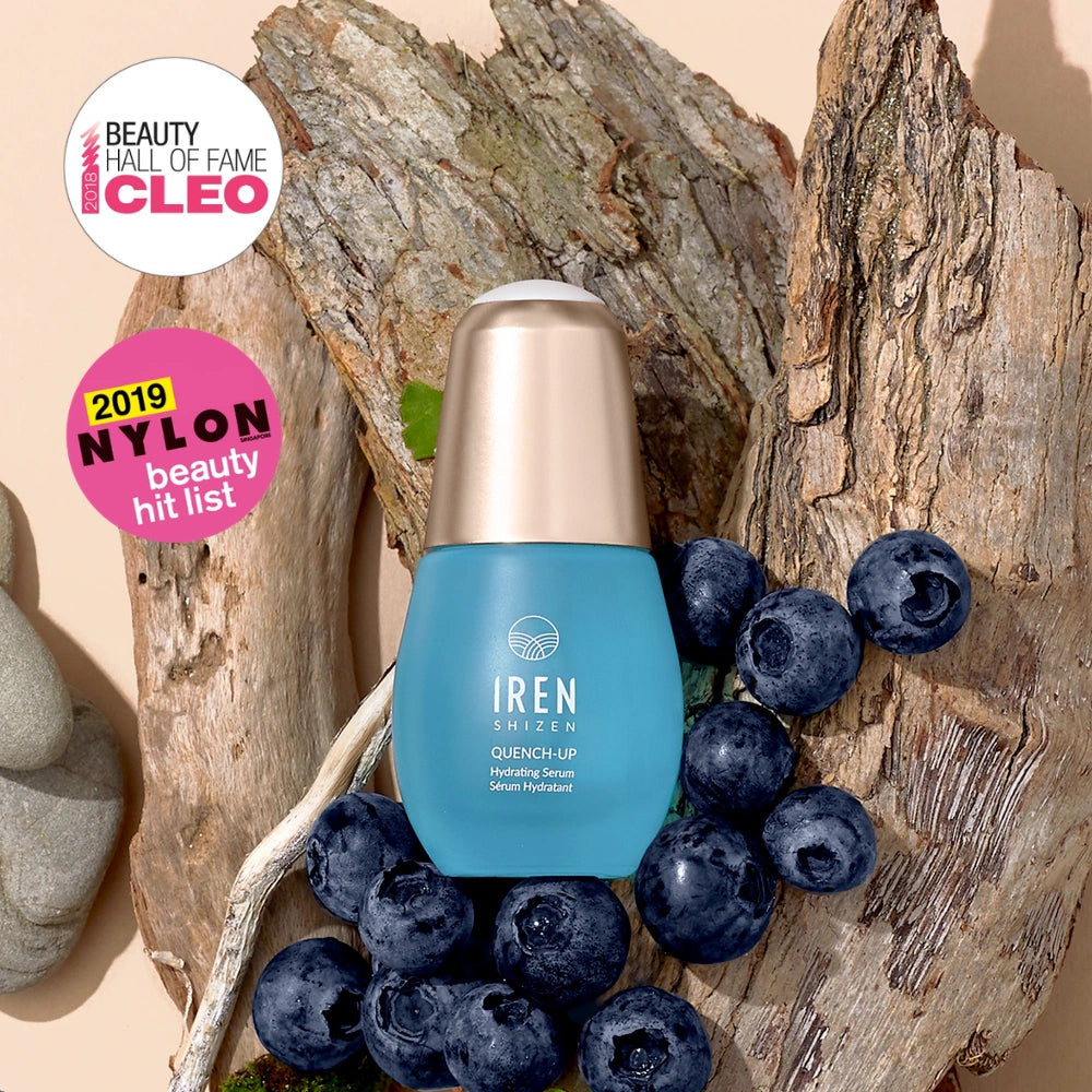 A blue bottle of IREN Shizen QUENCH-UP Hydrating Serum with hyaluronic acid is displayed on tree bark with blueberries, next to CLEO and NYLON beauty award badges.