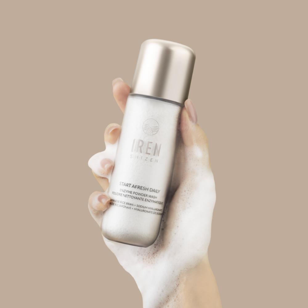 A hand holds a bottle from the FRESH AND CLEAR Cleansing Kit by Fast Bundle, covered in foamy lather, against a plain beige background—perfect for your skincare routine.