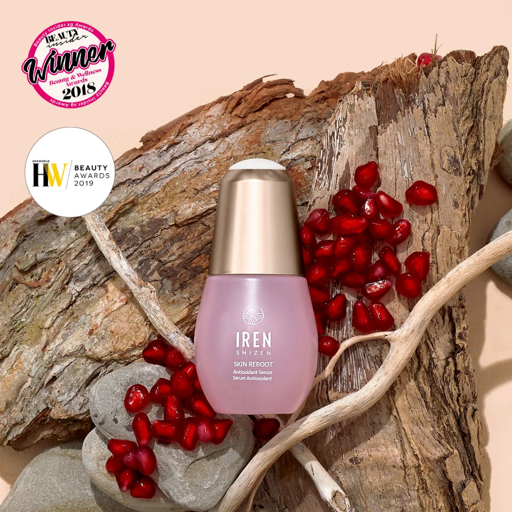 A bottle of IREN Shizen SKIN REBOOT Antioxidant Serum rests on wood with pomegranate seeds, rocks, and two beauty award badges, emphasizing its pomegranate extract for reducing dark circles. #all-groups