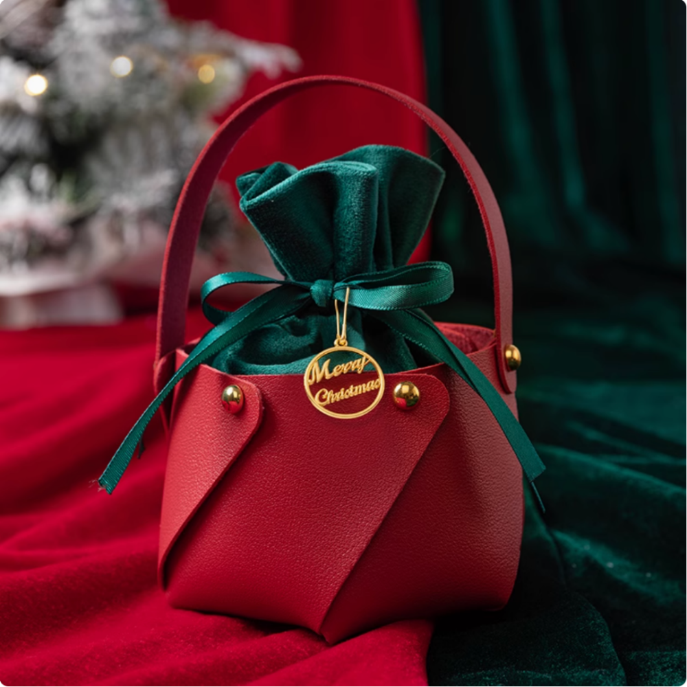 The IREN Shizen FOR THE PALS Xmas Apple Gift Bag features a red leather pouch adorned with gold studs, which holds a green velvet bag tied with a ribbon. A gold "Merry Christmas" tag is attached to the ribbon, while holiday decor adds to the festive backdrop.