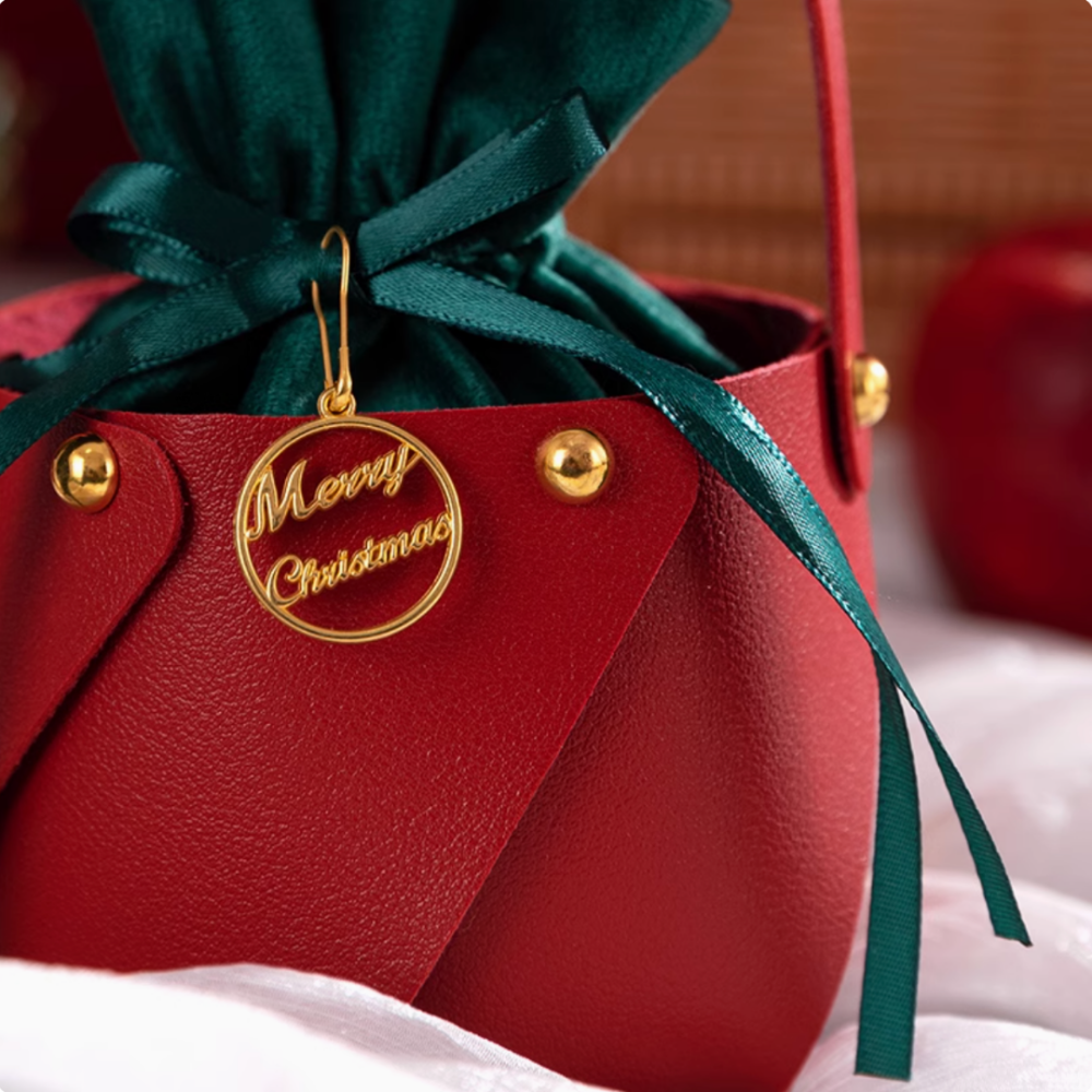 FOR THE PALS Xmas Apple Gift Bag by IREN Shizen features red leather with gold accents and a green ribbon adorning the top, complete with a gold circular "Merry Christmas" tag.