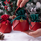 Hands holding an "IREN Shizen FOR THE PALS Xmas Apple Gift Bag" adorned with a "Merry Christmas" tag, surrounded by similar gift bags on a snowy surface with pine cones and festive greenery in the background.