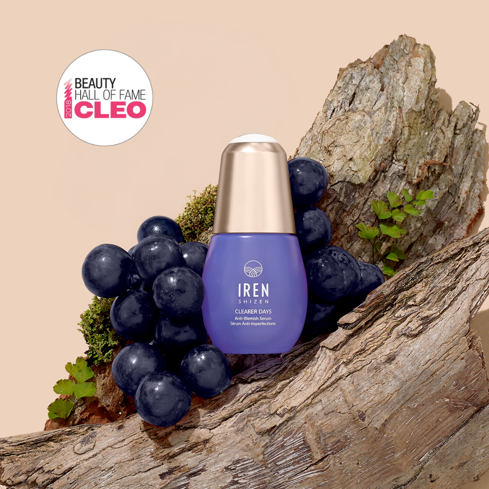 A bottle of IREN Shizen CLEARER DAYS Anti-Blemish Serum sits among grapes and tree bark, featuring a "Beauty Hall of Fame CLEO" badge in the top left corner.